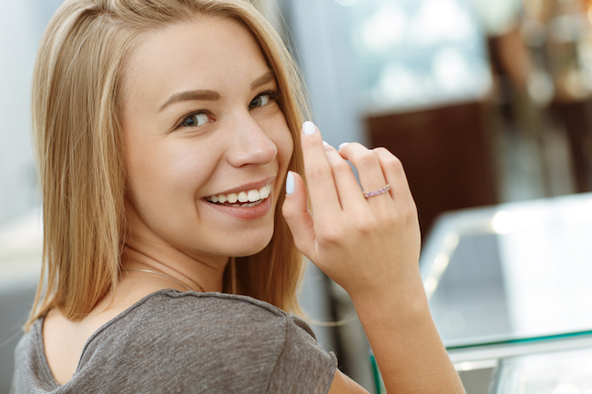 View from of attractive smiling woman looking at camera and posing in jewelry store. Pretty cheerful blonde trying on new accassories and showing shiny ring on hand. Concept of shopping.
